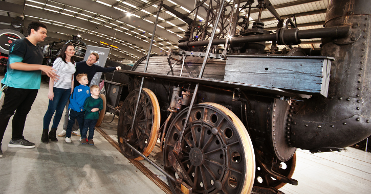 Family with children talking to a staff member stood next to an historic railway engine at Locomotion museum.